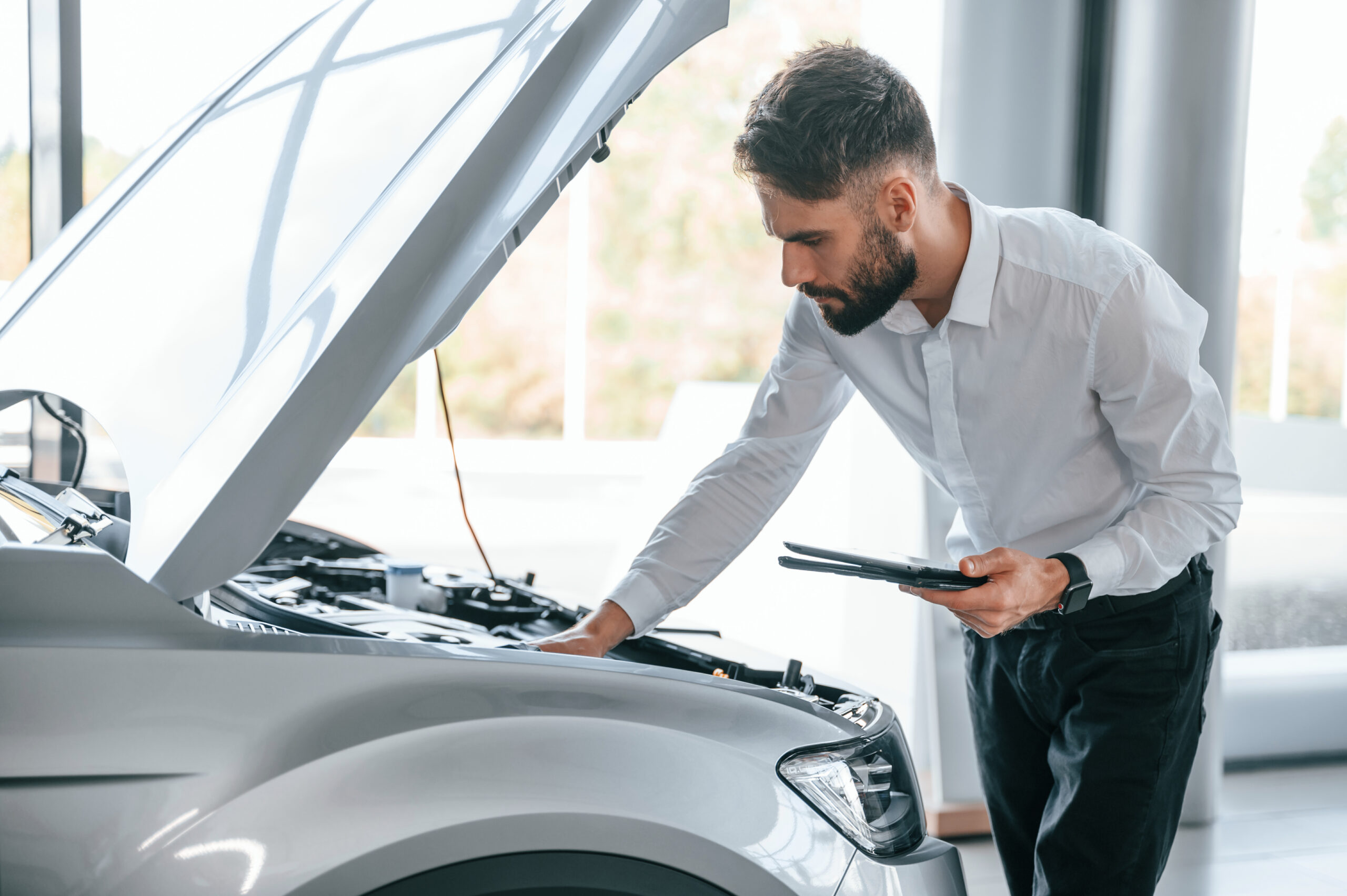 Looks Under The Hood Of Automobile. Young Man In White Clothes Is In The Car Dealership